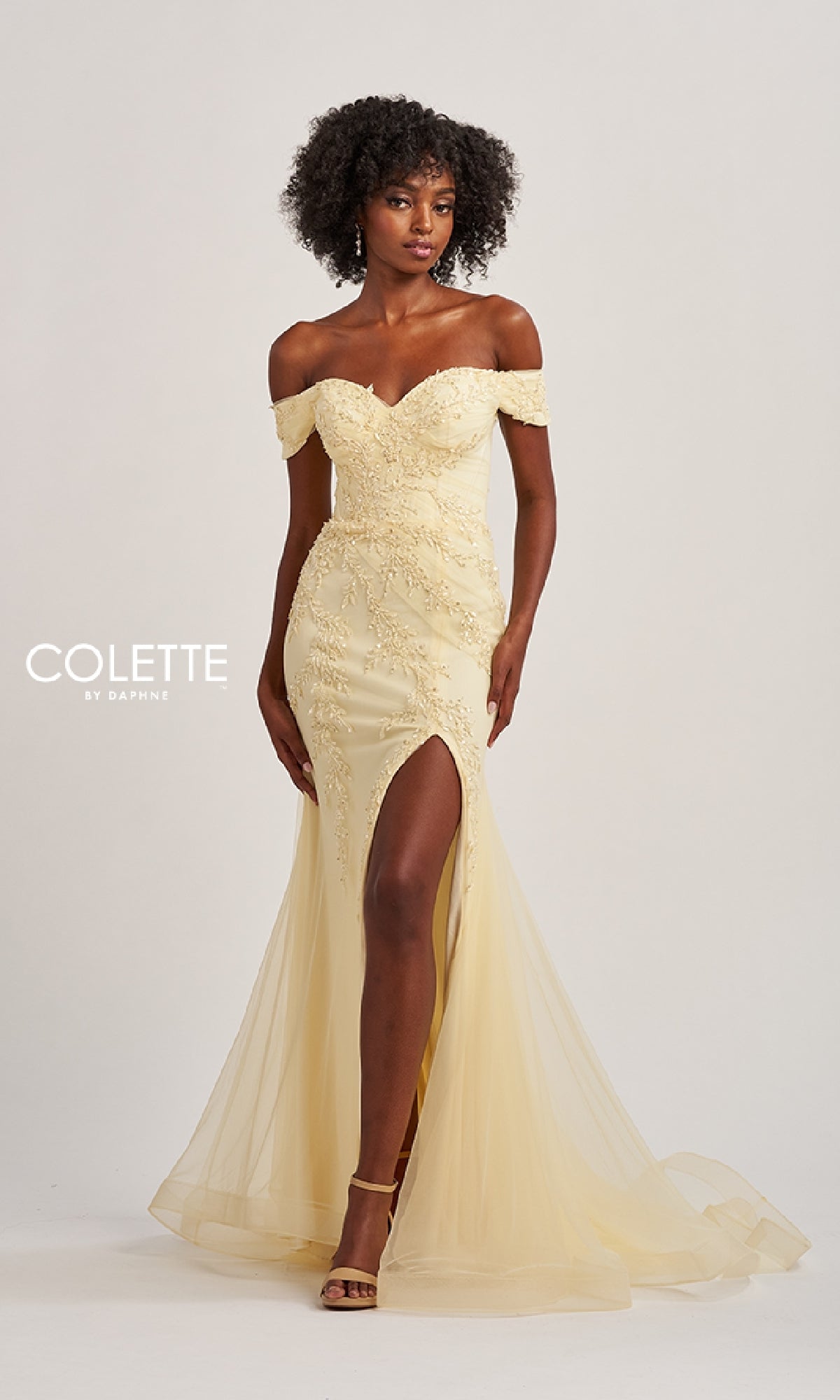 A woman in a pale yellow Off-the-Shoulder Long Prom Dress: Colette CL8645, featuring lace details and a thigh-high slit, stands against a plain background.